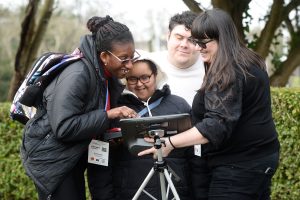 Young Lady tapping on a AAC device while another young lady watches and man looks over sholder