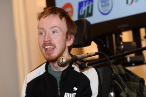 Speaker Marcus in a wheelchair beside a  microphone at the CHAT “AT in Action: From Rights to Reality” event, standing in front of a presentation screen displaying the event title and date
