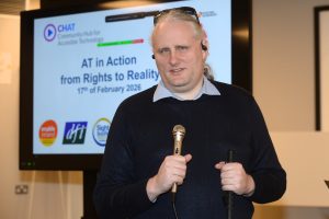 Speaker Declan Meenagh holding a microphone at the CHAT “AT in Action: From Rights to Reality” event, standing in front of a presentation screen displaying the event title and date