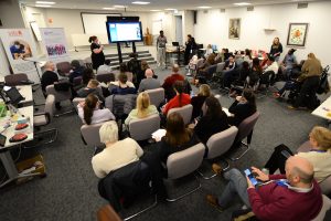 Wide view of attendees seated in a conference room during the CHAT “AT in Action” event, listening to a speaker presenting at the front beside a large screen.