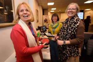 Three women smiling at a CHAT Assistive Technology event, standing beside a table displaying assistive devices, including a large red button switch, in a conference room setting.