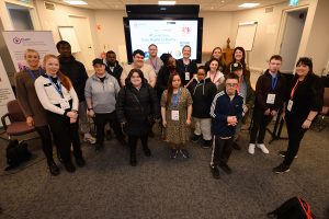 Group of attendees at a CHAT Assistive Technology event standing together in a conference room in front of a presentation screen, wearing name badges and facing the camera