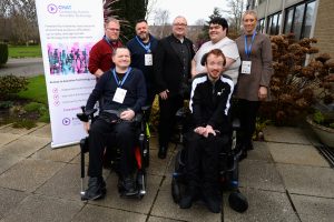 Group of eight adults at a CHAT Assistive Technology event standing outdoors beside a branded banner, with two wheelchair users positioned at the front and others standing behind, all wearing event name badges.